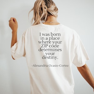 May include: Cream-colored t-shirt with the quote "I was born in a place where your ZIP code determines your destiny." attributed to Alexandria Ocasio-Cortez. The text is printed in black, centered on the back of the shirt.