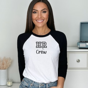 Op de afbeelding: Een wit en zwart baseballshirt met de tekst "HR Crew" in zwarte letters. Het shirt heeft een zwarte kraag en zwarte mouwen.