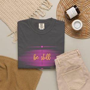 May include: A dark gray t-shirt with the words "be still" in a cursive font, set against a pink striped design. The shirt also features a small pink heart and the word "PSALM" at the bottom. The shirt is folded on a neutral surface.