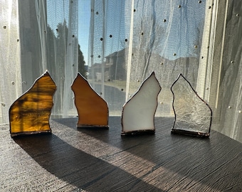 May include: Four cat-shaped stained glass ornaments with copper foil edges. The first is amber with dark streaks, the second is solid light orange, the third is opaque white, and the fourth is clear with a textured, crinkled glass surface.