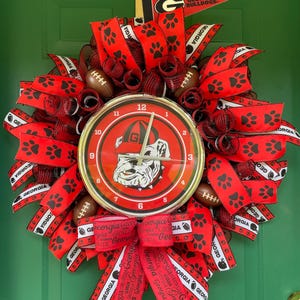 May include: A decorative wreath featuring red and black mesh, ribbons with paw prints and the text Georgia, and small footballs. A round clock face is centered, displaying a bulldog wearing a cap with a G logo and the text Georgia Bulldogs.