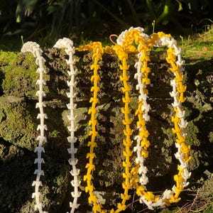May include: Four leis, traditional Hawaiian necklaces, are displayed. Two are white, one is yellow, and one is a combination of white and yellow. The leis are made of small, repeating, leaf-shaped elements, and are draped over a mossy rock.