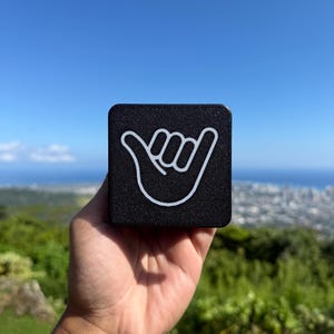 May include: A square black object with a textured surface featuring a white line art graphic of a shaka hand gesture. The object has rounded corners and a flat front face, displaying the iconic hand sign clearly against the dark background.
