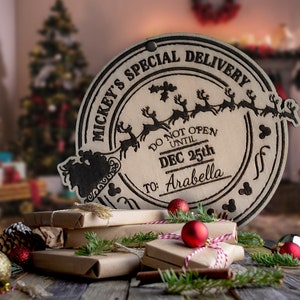 Op de afbeelding: Houten kerstdecoratie met de tekst "Mickey's Special Delivery" en "Do Not Open Until Dec 25th To: Arabella". De decoratie toont een silhouet van de slee van de kerstman en rendieren. Verschillende ingepakte cadeaus, ornamenten en groen staan op de voorgrond.