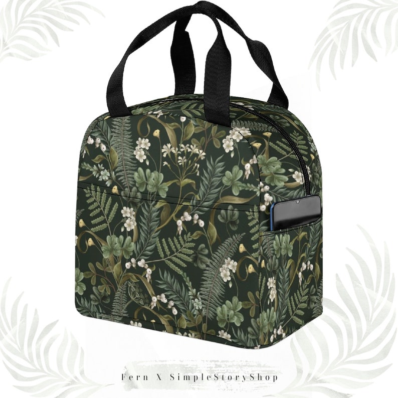 May include: A dark green insulated lunch bag with a floral pattern featuring white flowers and green foliage. The bag has a black zipper, black handles, and a pocket with a phone inside. The text "Fern X SimpleStoryShop" is at the bottom.