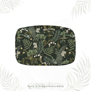 May include: A rectangular, dark green cosmetic bag with a botanical pattern. The pattern features ferns, white flowers, and berries in shades of green, white, and brown. The bag has rounded corners and a zipper closure.
