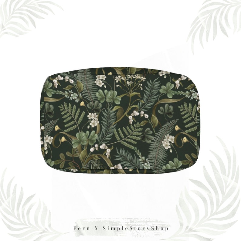 May include: A rectangular, dark green cosmetic bag with a botanical pattern. The pattern features ferns, white flowers, and berries in shades of green, white, and brown. The bag has rounded corners and a zipper closure.