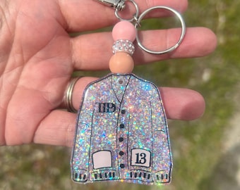 May include: A silver glitter keychain shaped like a cardigan sweater with a pink and white beaded charm. The sweater has a pocket on the front and the numbers "13" and "11" on the front.