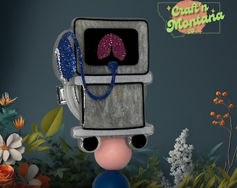 May include: A silver glitter resin brooch shaped like a medical breathing machine with a pink glitter heart on the screen. The brooch is on a white pedestal with a pink and blue ball on top.