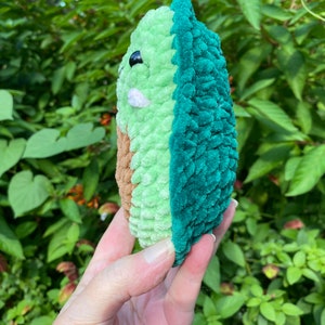 May include: A crocheted avocado plush toy with a green and brown exterior. The avocado is held in a hand with the pit facing the viewer.