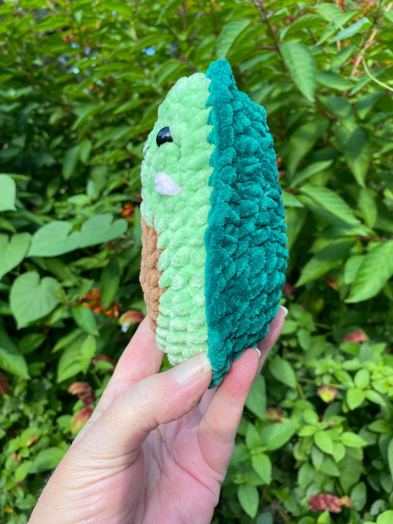 May include: A crocheted avocado plush toy with a green and brown exterior. The avocado is held in a hand with the pit facing the viewer.
