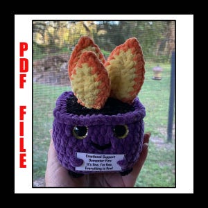 Könnte beinhalten: Ein gehäkelter lila Topf mit einem lächelnden Gesicht und zwei orangefarbenen und gelben Flammen, die aus der Oberseite herausragen. Der Topf hat ein Schild mit der Aufschrift "Emotional Support Dumpster Fire It's fine, I'm fine. Everything is final!"