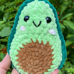 May include: A green and brown crocheted avocado plush toy with a cute face. The avocado has a dark green outer layer and a brown pit.