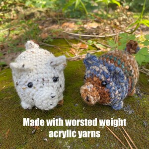 Op de afbeelding: Twee gehaakte varkensbeeldjes. E&eacute;n is wit met zwarte spikkels, de andere is bruin, blauw en bruin. Beide hebben zwarte ogen en kleine oren. De tekst "Made with worsted weight acrylic yarns" staat onderaan.
