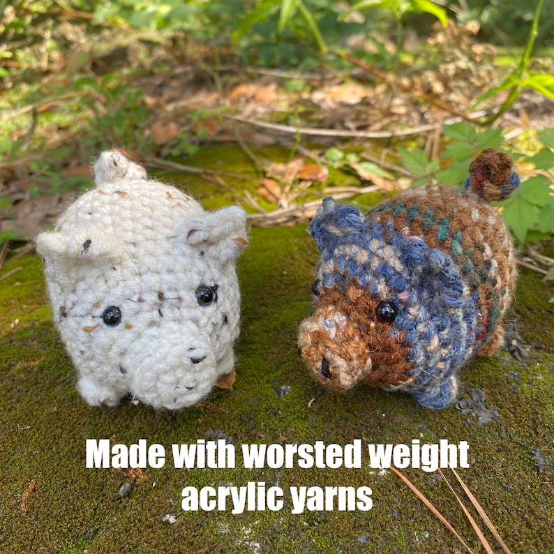 Op de afbeelding: Twee gehaakte varkensbeeldjes. E&eacute;n is wit met zwarte spikkels, de andere is bruin, blauw en bruin. Beide hebben zwarte ogen en kleine oren. De tekst "Made with worsted weight acrylic yarns" staat onderaan.