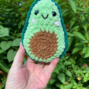 May include: A green crocheted avocado plush toy with a brown seed and a smiling face. The toy has black eyes and a white stitched smile.