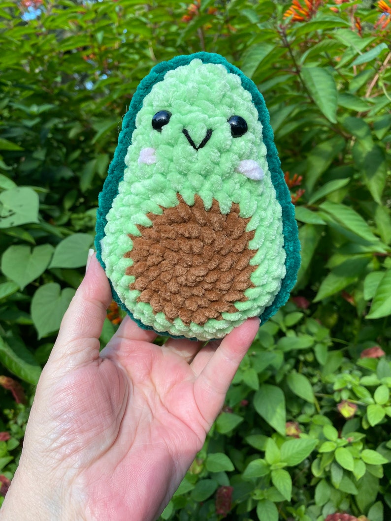 May include: A green crocheted avocado plush toy with a brown seed and a smiling face. The toy has black eyes and a white stitched smile.