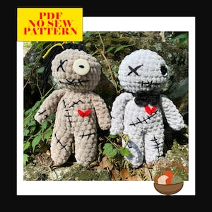 May include: Two crocheted voodoo dolls with stitched-on details, one with a black bow tie and one with a single button eye. The dolls are sitting on a bed of green leaves and rocks.