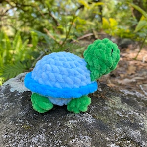 Pode incluir: Um brinquedo de tartaruga de croch&ecirc; azul e verde. A tartaruga tem uma carapa&ccedil;a azul claro com uma borda azul e uma cabe&ccedil;a e pernas verdes.