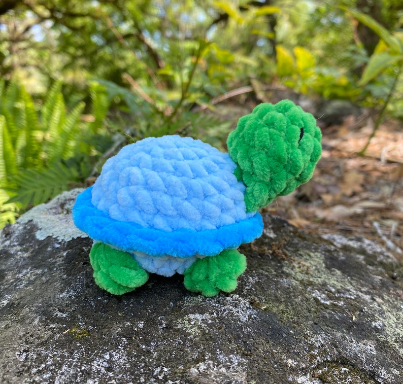 Pode incluir: Um brinquedo de tartaruga de croch&ecirc; azul e verde. A tartaruga tem uma carapa&ccedil;a azul claro com uma borda azul e uma cabe&ccedil;a e pernas verdes.