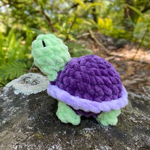 Pode incluir: Um brinquedo de tartaruga de croch&ecirc; com uma casca verde e detalhes roxos e lavanda.