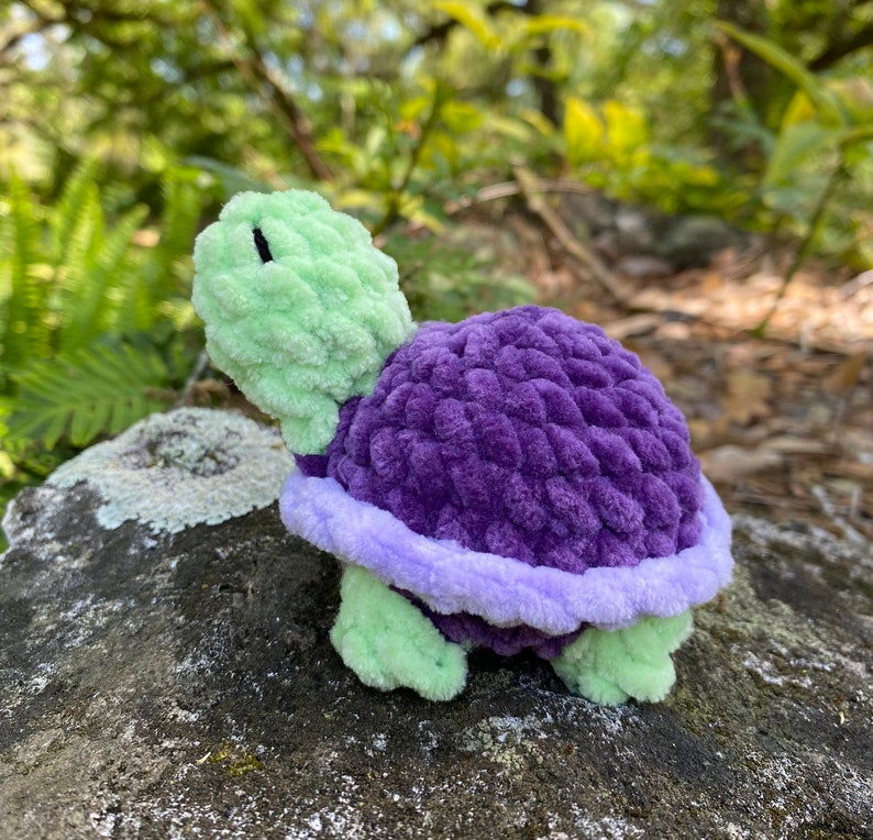 Pode incluir: Um brinquedo de tartaruga de croch&ecirc; com uma casca verde e detalhes roxos e lavanda.