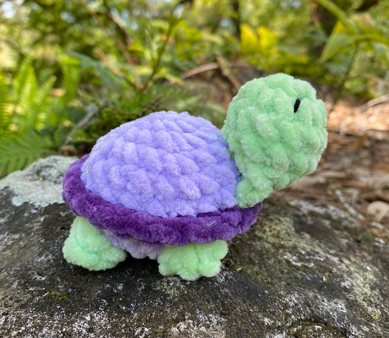 Pode incluir: Uma tartaruga de pel&uacute;cia com uma concha roxa, cabe&ccedil;a e pernas verdes. A concha tem um design texturizado e croch&ecirc;.