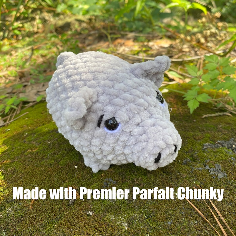 Op de afbeelding: Een handgemaakt, grijs gehaakt varkentje met zwarte ogen en een zwarte snuit. Het varken is gemaakt met Premier Parfait Chunky garen en staat op een mosachtig oppervlak. De tekst "Made with Premier Parfait Chunky" is zichtbaar.