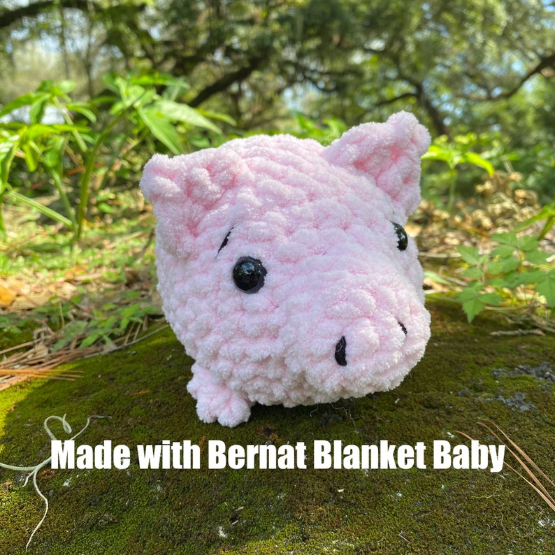 Op de afbeelding: Een handgemaakt, lichtroze gehaakt varken met zwarte kraaloogjes en een kleine snuit. Het varken is gemaakt met Bernat Blanket Baby garen. Het varken zit op mos. De tekst "Made with Bernat Blanket Baby" is zichtbaar.