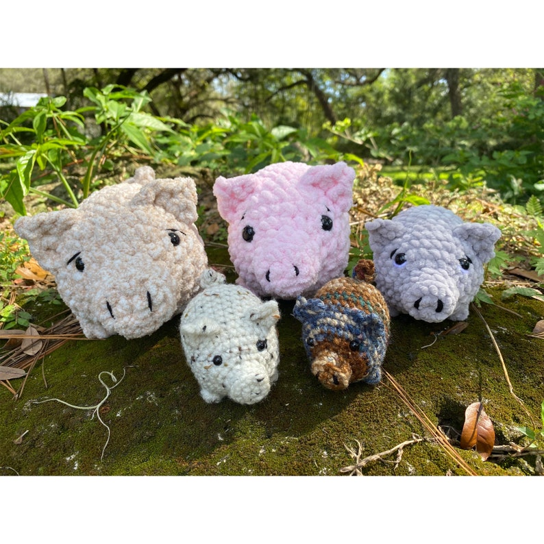 Op de afbeelding: Een verzameling van vijf gehaakte varkensbeeldjes in verschillende kleuren, waaronder roze, grijs, beige en blauw. De varkens hebben kleine, zwarte ogen en zijn gerangschikt op een mosachtig oppervlak, met een natuurlijke achtergrond van groen gebladerte.