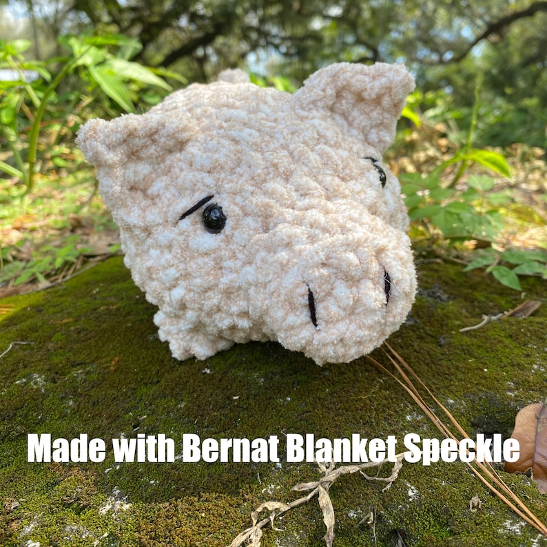 Op de afbeelding: Een zacht, beige varkenshoofd gemaakt met Bernat Blanket Speckle garen. Het varken heeft zwarte ogen en een snuit. De foto is buiten genomen, met groene bladeren op de achtergrond.