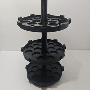 May include: A black, three-tiered display stand with a central pole and circular trays. Each tray has multiple semi-circular cutouts. The stand is made of a matte black material and is designed to hold small items.