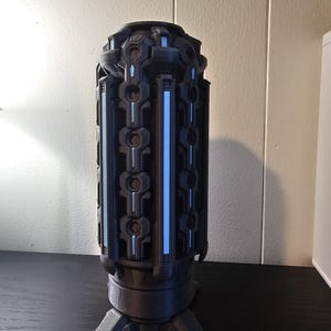 May include: A cylindrical dark gray object with a complex mechanical texture and vertical light blue accents. The structure features a series of repeating geometric patterns along its length and stands on a base with three short, angled legs.