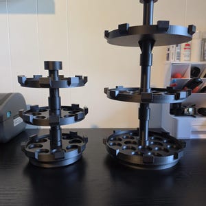 May include: Two black, tiered, circular storage organizers with multiple round slots for holding small items. The shorter organizer has three levels, while the taller organizer features three levels with a central vertical support post for each.