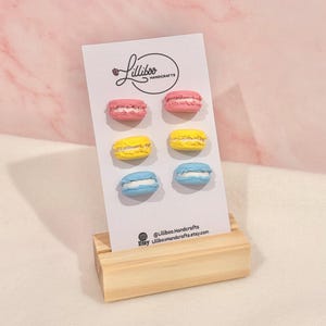 Könnte beinhalten: Präsentation von sechs bunten Ohrringen in Macaron-Form auf einer weißen Karte, gehalten von einem Holzständer. Die Ohrringe sind rosa, gelb und blau, jeweils mit einer weißen Füllung. Die Karte zeigt das Logo "Lilliboo Handcrafts".