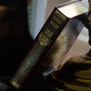May include: A close-up of a vintage book titled "Nicholas Nickleby" by Charles Dickens, with gold lettering and accents. The book is dark red and resting next to a brass candlestick holder.