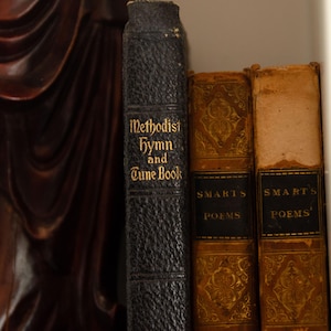 May include: A collection of antique books on a shelf. The books include a black leather-bound "Methodist Hymn and Tune Book" and two volumes of "Smart's Poems" with gold embossed covers. The books are in varying states of wear.