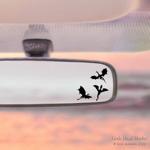 May include: Black dragon decals are affixed to a car's rearview mirror. The decals are in flight, with wings spread. The background is a soft, pastel sky. The text "Little Decal Market" and "little moments of joy" is in the lower right corner.