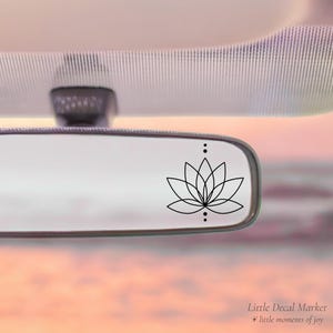 May include: A black lotus flower decal is affixed to a car's rearview mirror. The decal features a minimalist design with delicate lines and three small dots. The background shows a soft, pastel-colored sky, and the text "Little Decal Market" is visible.
