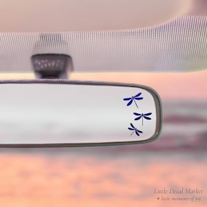 May include: A car's rearview mirror with three dark blue dragonfly decals. The decals are arranged vertically, decreasing in size. The background is a soft, blurred gradient of pink and orange.