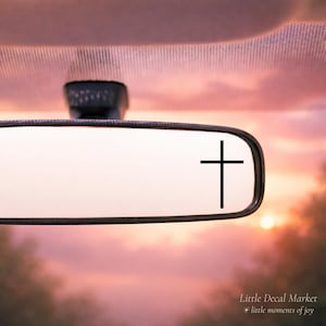 May include: A car's rearview mirror with a black cross decal. The background shows a sunset with pink and orange hues. The text "Little Decal Market + little moments of joy" is at the bottom.