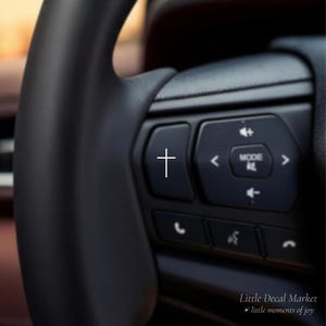 May include: Close-up of a black steering wheel with integrated control buttons. A white cross symbol is on one button. Other buttons include volume, mode, and phone controls. The text "Little Decal Market + little moments of joy" is at the bottom.