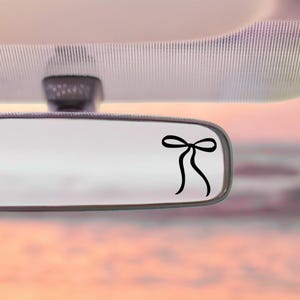 May include: A black bow decal is affixed to a car's rearview mirror. The bow is a simple, elegant design with two loops and flowing ribbons. The mirror reflects a soft, blurred background.