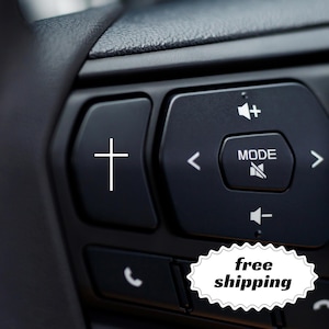 May include: Close-up of a black car steering wheel control panel. The panel features buttons with white symbols, including a cross, volume controls, and a "MODE" button. The text "free shipping" is also visible.