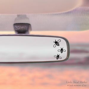 May include: A car's rearview mirror with three black bee decals. The bees are in a vertical arrangement, with the top bee slightly larger than the other two. The background is a soft, blurred sunset.
