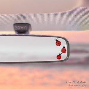 May include: A car's rearview mirror with three red ladybug decals. The ladybugs have black spots and antennae. The background is a soft, pastel sky. The text "Little Decal Market" and "little moments of joy" is in the bottom right corner.