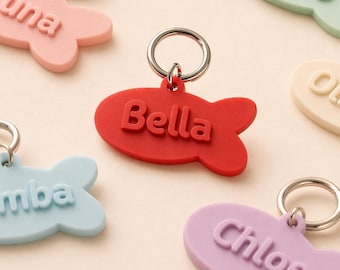Può includere: Una collezione di targhette identificative per animali domestici a forma di pesce, colorate in pastello, con nomi in rilievo come "Luna" e "Bella". Ogni targhetta è di un colore pastello diverso, tra cui rosa, rosso, blu, verde e viola, e ha un portachiavi argentato.