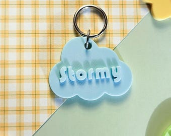Può includere: Un portachiavi a forma di nuvola azzurra con il nome "Stormy" in lettere bianche in rilievo. Il portachiavi ha un anello argentato ed è posto su uno sfondo a scacchi giallo e bianco con altre forme colorate.