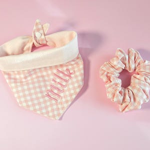 May include: A pink and white gingham dog bandana with the embroidered word "Milk" and a matching scrunchie. The bandana has a light pink trim and a button closure. The scrunchie is made of the same gingham fabric.