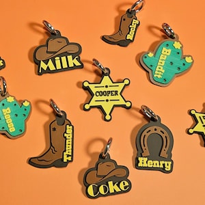 Könnte beinhalten: Eine Sammlung von Schlüsselanhängern im Western-Stil mit verschiedenen Designs, darunter ein Hufeisen, ein Cowboyhut, ein Kaktus, ein Sheriff-Abzeichen und ein Cowboy-Stiefel. Jeder Schlüsselanhänger hat einen silbernen Ring und einen Namen wie "Oliver", "Milk", "Rocky" und "Bandit".